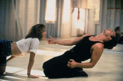 Patrick Swayze as Johnny Castle, a light-skinned man with short, styled brown hair wearing a black tank top and black pants is kneeling on the ground of a dance studio playing air guitar. Next to him is Jennifer Grey as Baby, a petite, light-skinned women with medium-long, wavy brown hair wearing a cropped white T-shirt and jeans shorts. Baby is crawling on her hands and knees toward Johnny. 